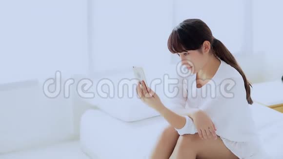 美丽的肖像年轻的亚洲女人坐在卧室里用智能手机自拍早上在家视频的预览图