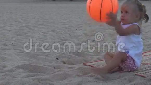 在海滩上穿着白色T恤的漂亮宝宝朝那个方向扔了一个橙色的大球视频的预览图
