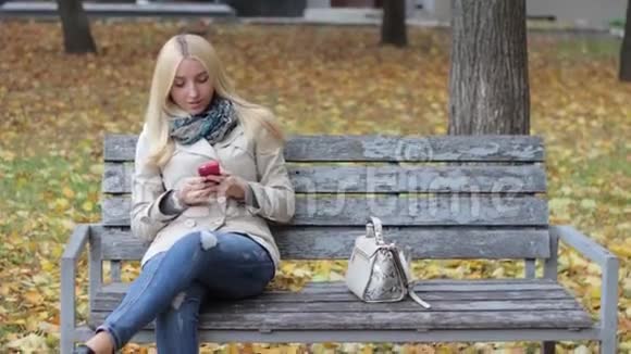 一个年轻漂亮的金发女孩坐在秋天公园的长凳上手里拿着手机视频的预览图