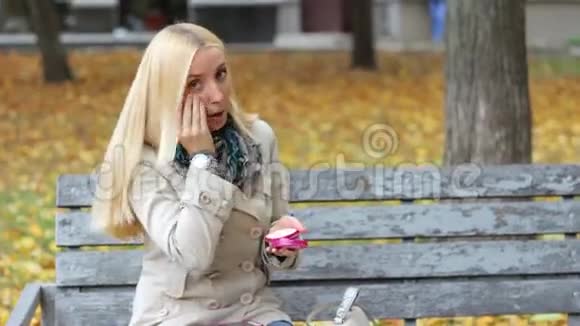 一个年轻漂亮的金发女孩坐在秋天公园的长凳上手里拿着手机视频的预览图