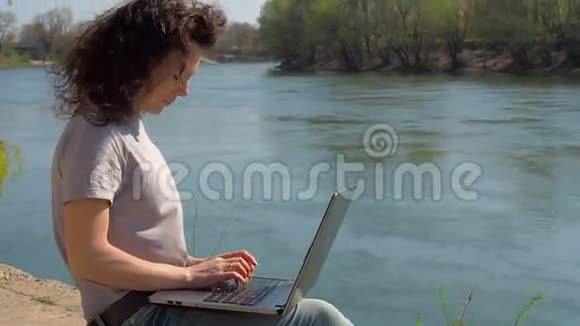 有笔记本电脑的女孩一个女人正在河边的笔记本电脑上工作一个春天阳光明媚的日子视频的预览图