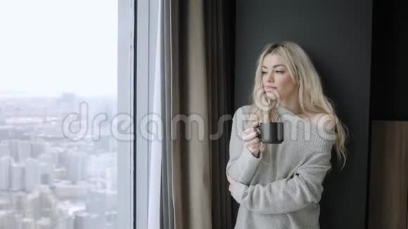 美丽的金发女人拿着陶瓷杯的茶或咖啡享受在床上喝茶看着窗户喝茶视频的预览图