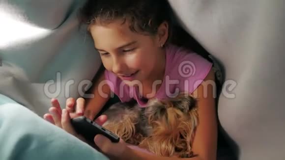 少女和狗在毯子下玩智能手机小女孩睡前躺在床上冲浪视频的预览图