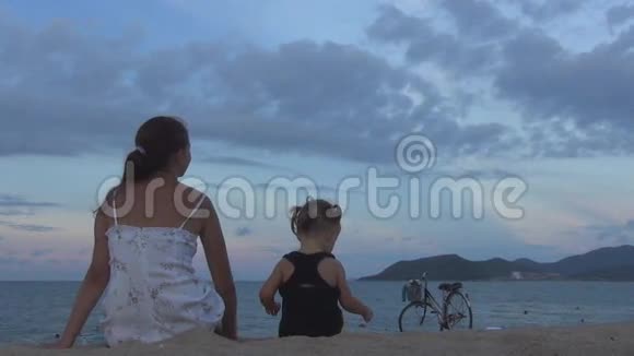美丽的女孩留着长发一个婴儿背对着沙滩上的摄像机坐着他们看着蓝天和一个视频的预览图