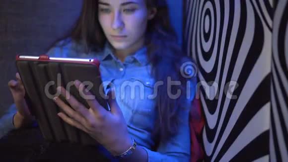 年轻漂亮的女孩坐在沙发上拿着平板电脑像斑马一样靠在墙上多种颜色的光相机视频的预览图