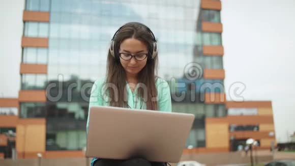 漂亮的年轻女孩带着耳机坐在空地上带着笔记本电脑视频的预览图