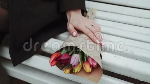 年轻女子坐在长凳上手捧郁金香花束特写天气寒冷白色视频的预览图