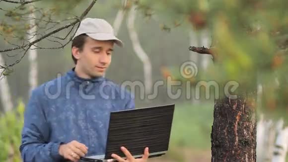 一个男人在森林里一棵树旁的笔记本电脑上打视频电话清晨视频的预览图