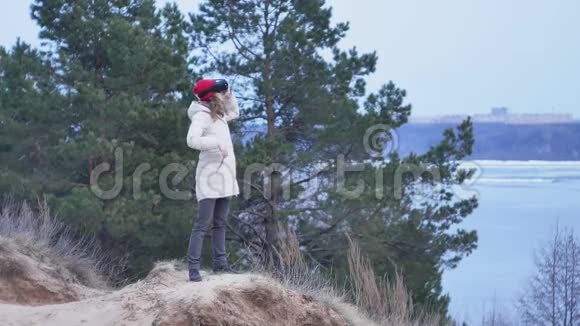 漂亮的年轻女游客戴着红帽子坐在岸上享受着虚拟现实头盔视频的预览图