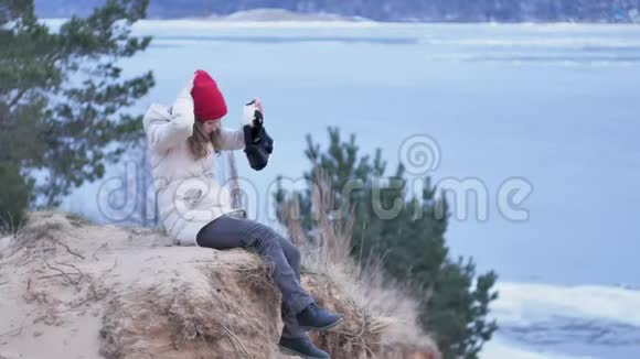 漂亮的年轻女游客戴着红帽子坐在岸上享受着虚拟现实头盔视频的预览图