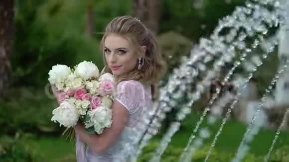 年轻的金发女郎穿着内衣在喷泉附近的花园里摆着新娘花束视频的预览图