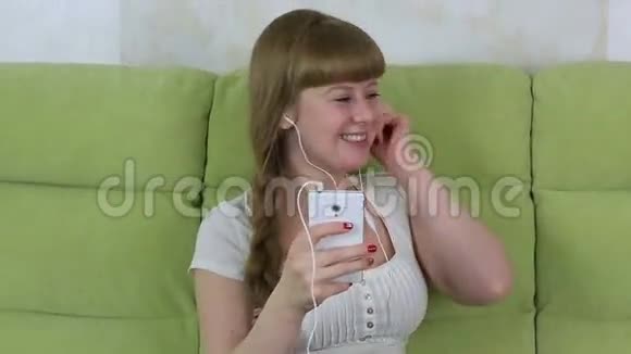年轻女孩坐在沙发上拿着智能手机用耳机听音乐视频的预览图