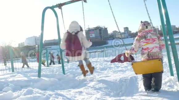 年轻的母亲带着孩子在冬季公园的户外秋千上摇摆降雪降雪冬季视频的预览图