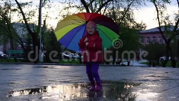 可爱的小女孩带着五颜六色的雨伞在日落时在公园的水坑里跳视频的预览图