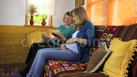 年轻女子抚摸怀孕的肚子和奶奶一起看书视频的预览图