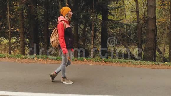一个年轻苗条的女孩戴着黄色的帽子和一个老式的背包在秋天的黄色针叶树上沿着沥青路散步视频的预览图
