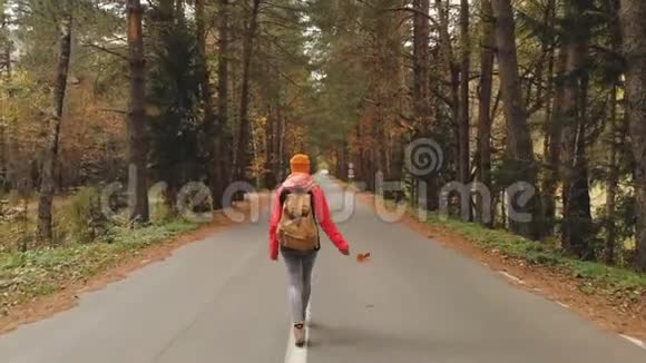 一个年轻苗条的女孩戴着黄色的帽子和一个老式的背包在秋天的黄色针叶树上沿着沥青路散步视频的预览图