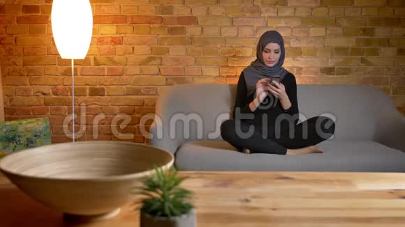 戴着头巾的穆斯林成年女性的特写镜头在舒适的室内沙发上坐着用电话视频的预览图