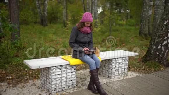 在秋天公园坐在长凳上时年轻的女人用平板电脑坐在长椅上的学生视频的预览图