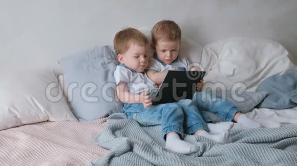 带平板电脑的孩子两个男孩双胞胎蹒跚学步看着躺在床上的平板电脑视频的预览图