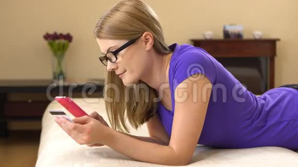 美丽迷人的年轻女人躺在沙发上带着智能手机和网上购物视频的预览图