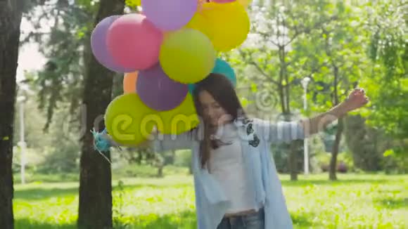 在阳光明媚的公园里散步时一个快乐顽皮的女孩摆着五颜六色的气球视频的预览图