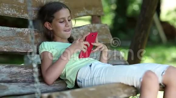 漂亮的女孩带着红色智能手机坐在花园里的秋千凳上视频的预览图