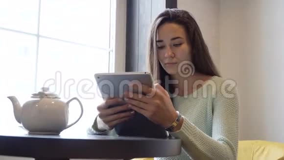 年轻的黑发女孩坐在靠近窗户的桌子上在平板电脑上工作视频的预览图
