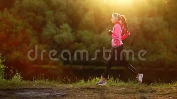 戴着长头发耳机的布鲁内特早上在公园里沿着河边跑步夏天的日出时穿着粉红色的衣服视频的预览图