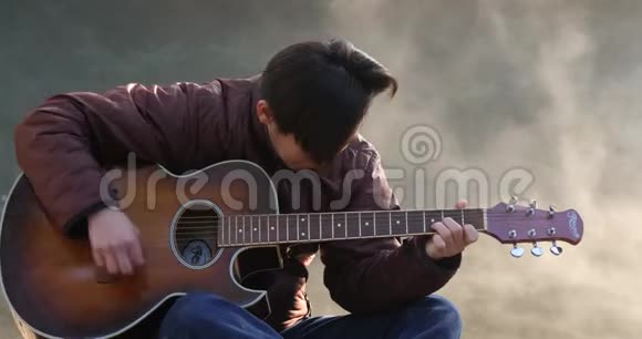 英俊的亚洲年轻人在日出时在森林河沿岸弹吉他黎明时分雾蒙蒙的背景视频的预览图