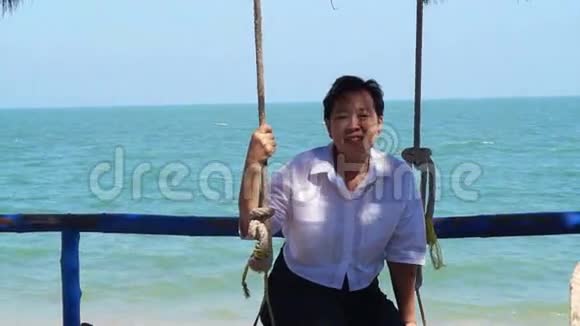 美丽的亚洲老年妇女在海滩上的秋千上放松蓝色的海洋背景视频的预览图