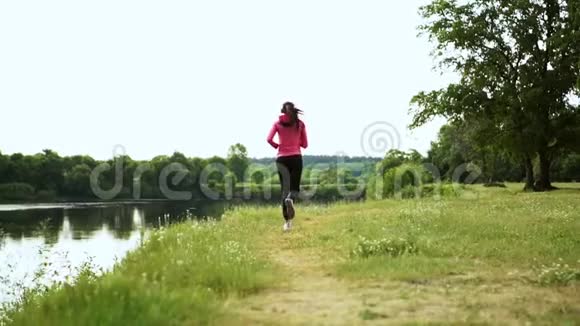 戴着长头发耳机的布鲁内特早上在公园里沿着河边跑步夏天的日出时穿着粉红色的衣服视频的预览图