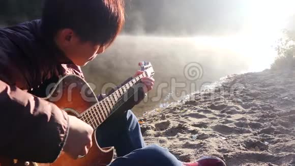 英俊的亚洲年轻人在日出时在森林河沿岸弹吉他黎明时分雾蒙蒙的背景视频的预览图
