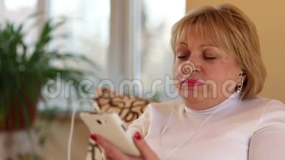 金发女人带着白色智能手机坐在沙发上听音乐视频的预览图