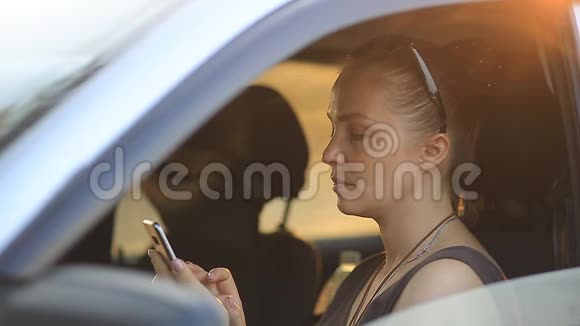 日落时分戴着墨镜红发的年轻美女坐在车轮后面手里拿着手机视频的预览图