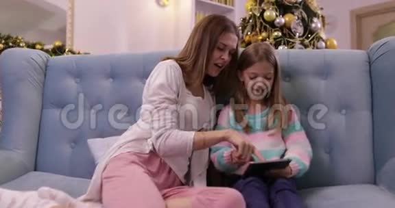 母亲和女儿在沙发上用平板电脑快乐微笑的年轻家庭附近装饰的新年圣诞树视频的预览图