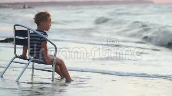 坐在海边椅子上的男孩海浪在洗他的衣服视频的预览图