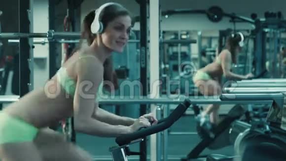 运动女性带着耳机在健身房的固定自行车上踩着模拟器视频的预览图