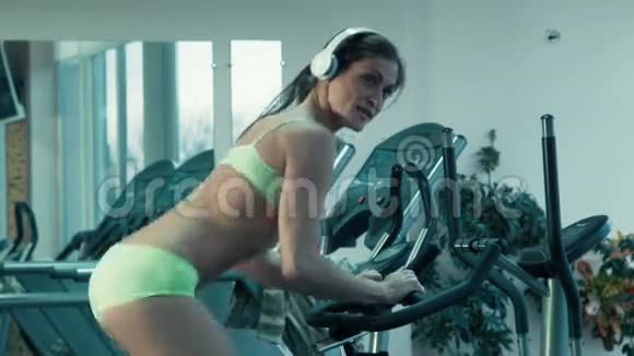 运动女性带着耳机在健身房的固定自行车上踩着模拟器视频的预览图