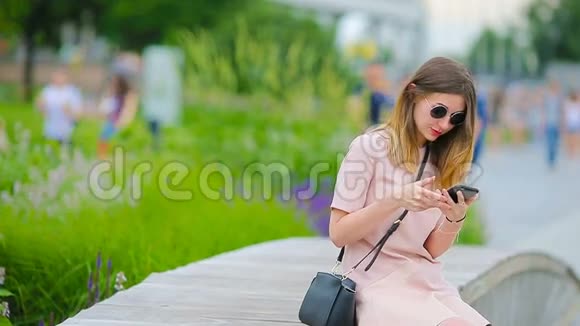 年轻的高加索女人在欧洲城市在户外发送信息和听音乐戴墨镜的漂亮女孩坐着视频的预览图
