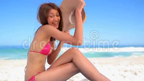 快乐的黑发女人戴着帽子坐在沙滩上视频的预览图