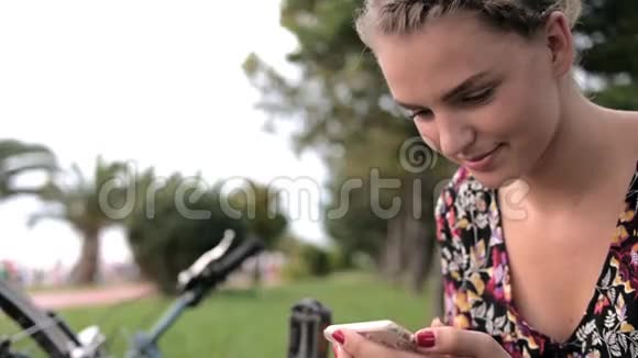 阳光明媚的一天一个漂亮可爱的女孩在公园里的自行车旁边用智能手机和手掌视频的预览图