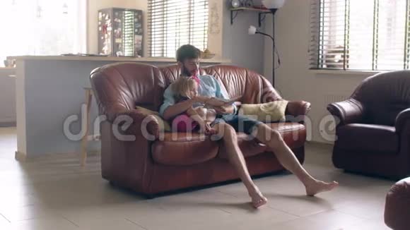 家庭幸福年轻的父亲和他的金发小女儿正在餐厅的皮革沙发上读一个故事4K视频的预览图