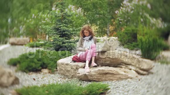 一个女人用耳机听音乐在一个美丽的绿色公园的岩石上放松一个常年年轻的女人视频的预览图