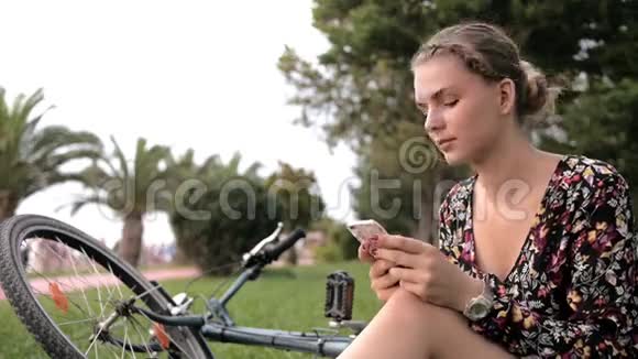 阳光明媚的一天一个漂亮可爱的女孩在公园里的自行车旁边用智能手机和手掌视频的预览图