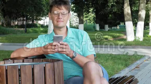 一个戴眼镜的人坐在公园的长凳上用电话视频的预览图