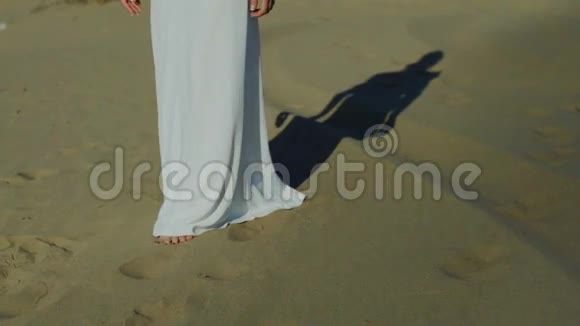 穿着白色连衣裙在沙滩沙丘上行走的女人的脚太阳落山时的影子慢动作视频的预览图