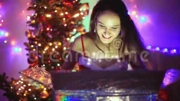 年轻快乐的女人坐在圣诞树旁边装饰着圣诞礼物和神奇的灯光视频的预览图