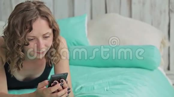 年轻的卷发女人躺在床上拿着智能手机多利射击视频的预览图