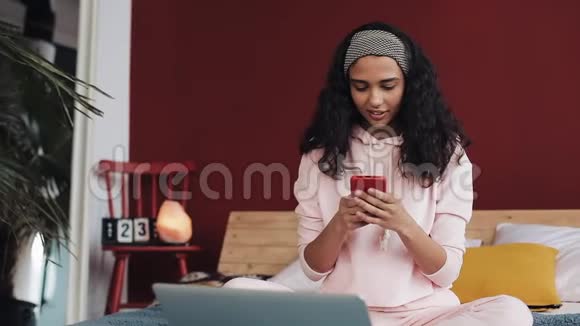 快乐的非裔美国女孩聊天和短信智能手机坐在床上在家里年轻女子视频的预览图